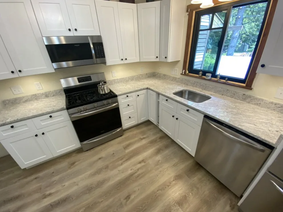 Wide view of a tailored kitchen renovation highlighting white shaker cabinets, stone countertops, and new wood-style flooring.