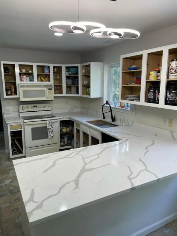 A wide-angle view of a kitchen in mid-renovation, displaying new white shaker cabinets and a significant Calacatta quartz installation on the L-shaped countertops and central peninsula, under modern ring lighting.