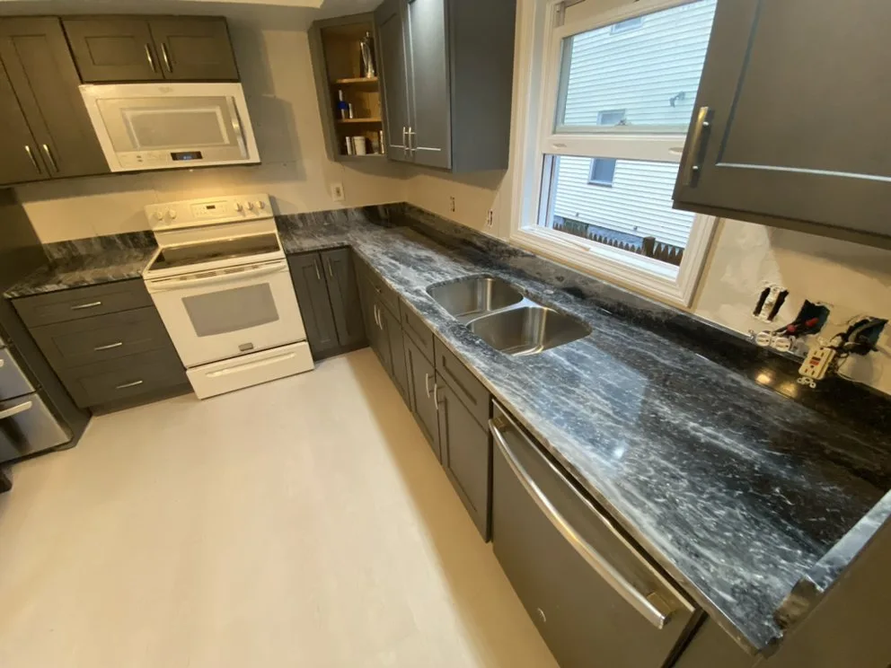 Completed granite countertop installation featuring dark veined surfaces, a stainless steel double sink, and grey cabinets.